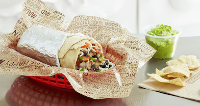 A burrito from the restaurant Chipotle, displayed in a red basket. To the right of the burrito is a side of chips and guacamole.