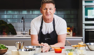 Chef Gordon Ramsay leaning on a kitchen counter, with silver mixing bowls to his left and ingredients in a bowl prepped for cooking to his right.