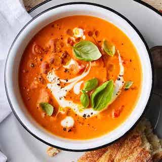 A white bowl filled with tomato soup. The soup is swirled with cream and topped with 7 pieces of fresh basil, all varying in sizes. To the bottom right of the bowl is a chunk of french bread.