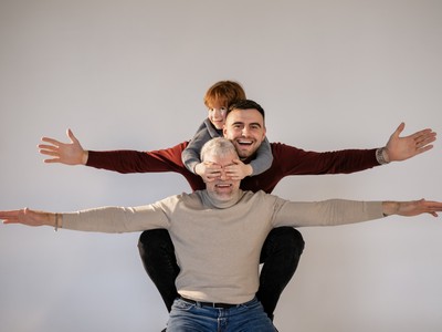 a boy sits on his father's back piggy-back style, and the father sits the same way on his dad's back. All three generations, with their arms sticking out to the sides as they smile. The boy covers his grandpa's eyes with his hands