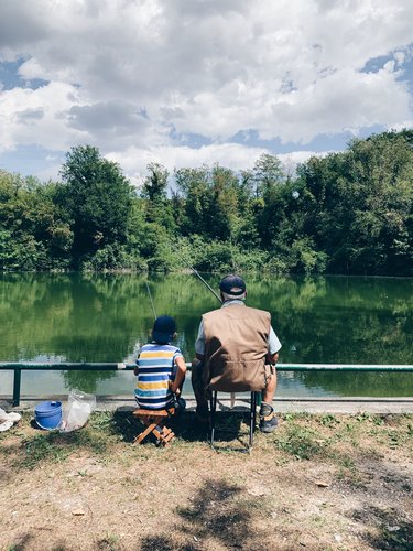 Older man and young boy sitting before a lake as they hold fishing poles and gaze at the water