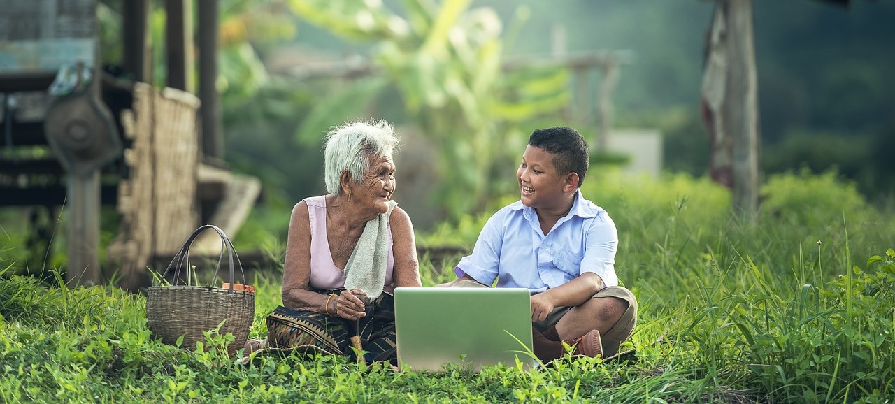 an old woman with white hair sits on the ground with a basket next to her. On her other side is a boy of about 12 who shows her his laptop. They sit on green grass and other short weeds, and behind them is a blurred image of a hut.