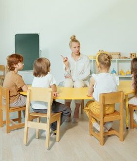 four elementary-age kids sit on wooden chairs as they listen to their teacher who sits across from them with her hair tied in a high bun