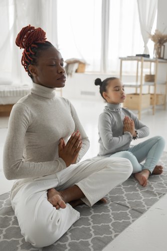 A woman with braided hair wrapped up in a high bun sits cross-legged on the floor in a meditation pose, a young girl with the same bun-style hair sits next to the woman and mimics her pose.