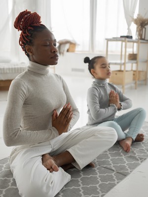 A woman with braided hair wrapped up in a high bun sits cross-legged on the floor in a meditation pose, a young girl with the same bun-style hair sits next to the woman and mimics her pose