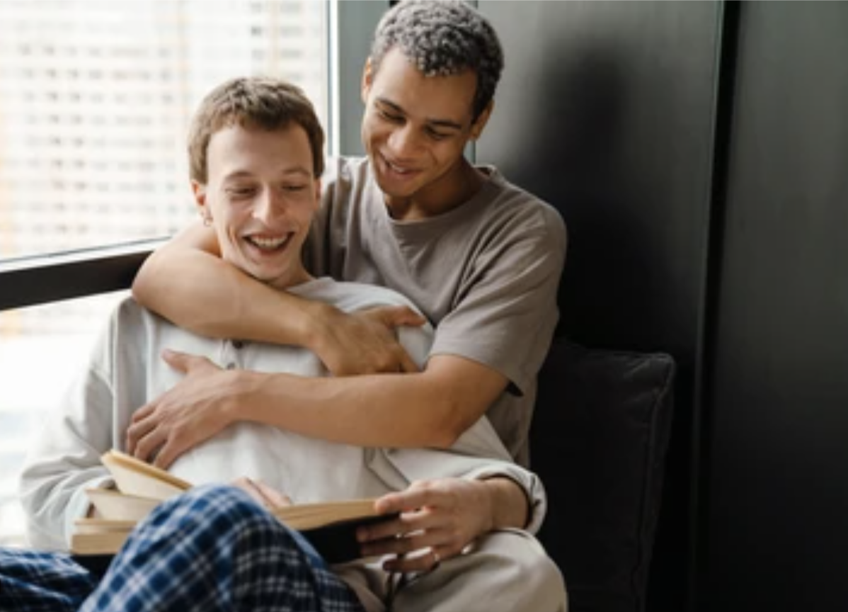 Man reads a book while his male partner sits behind with his arms wrapped around his front.