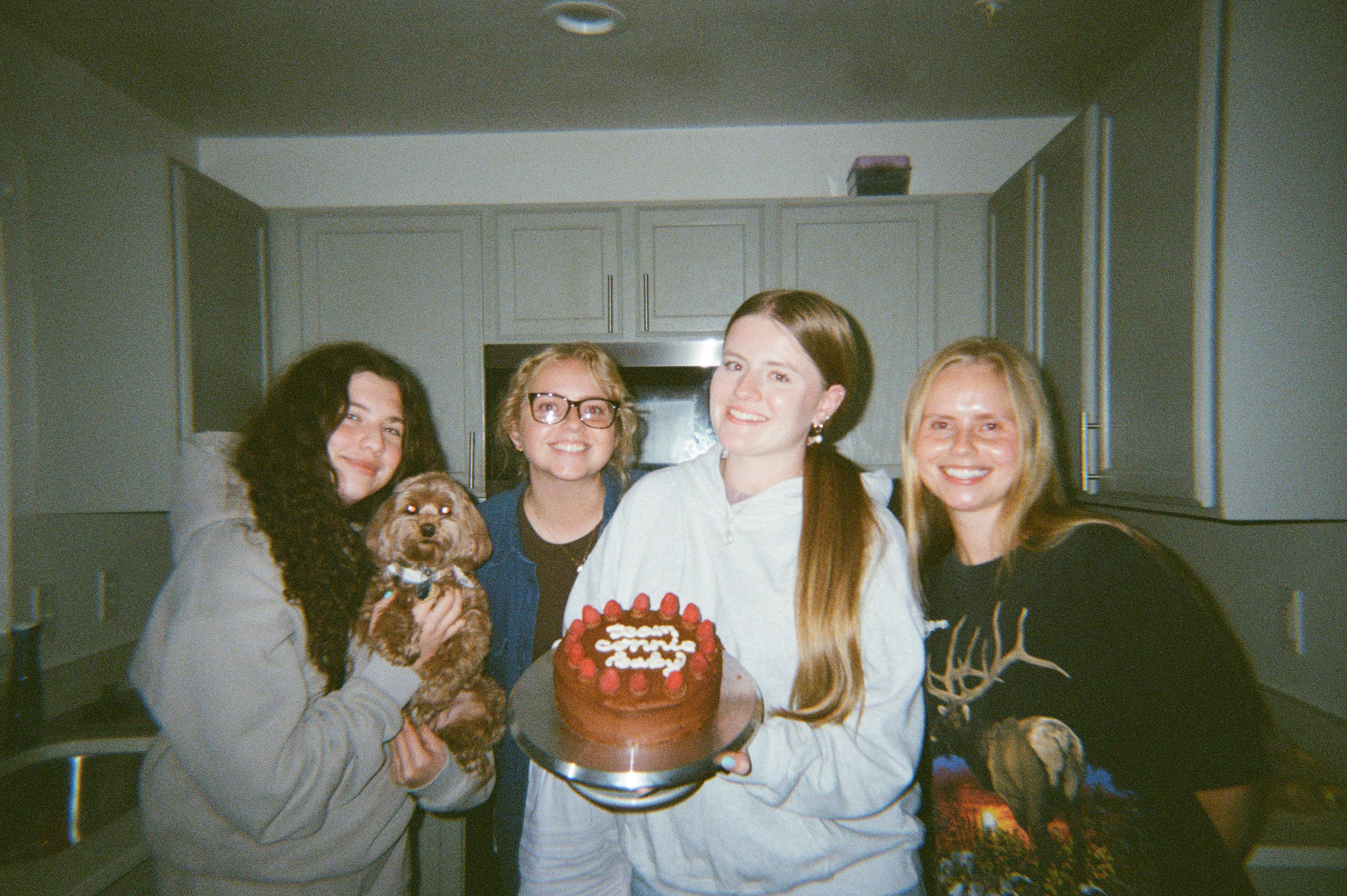 Four girls standing in a kitchen, one of which is holding a small dog, one of which is holding a chocolate cake with raspberries.