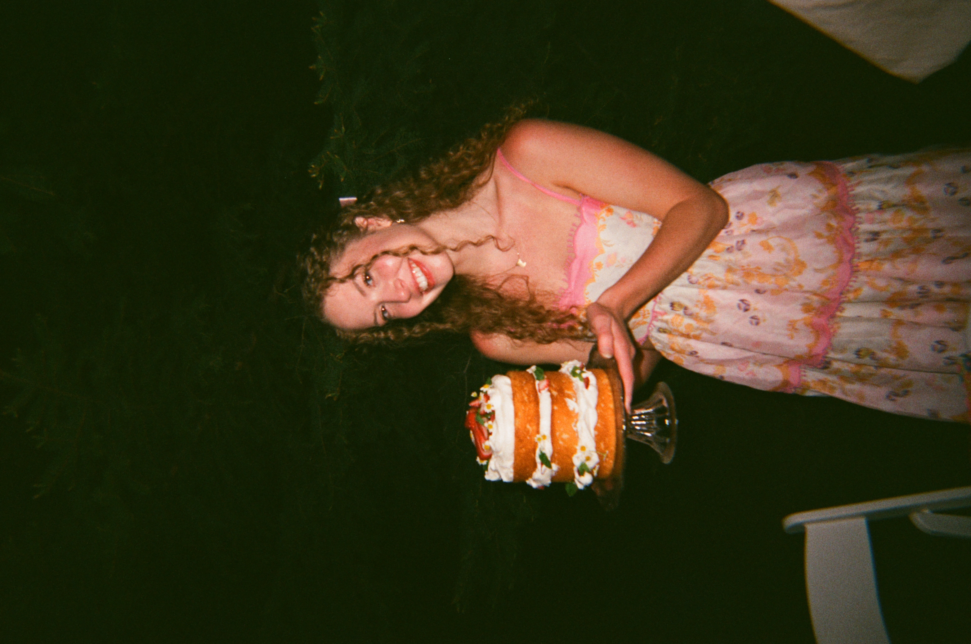 A young woman wearing a pink floral dress holding a cake.