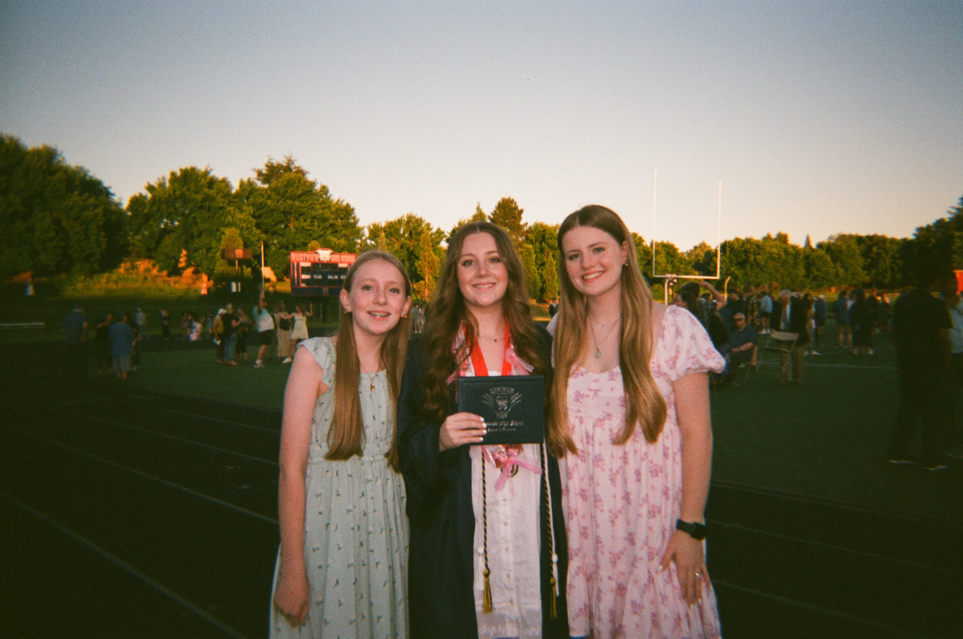 A young woman holding her diploma at high school graduation with one of her sisters on either side of her.