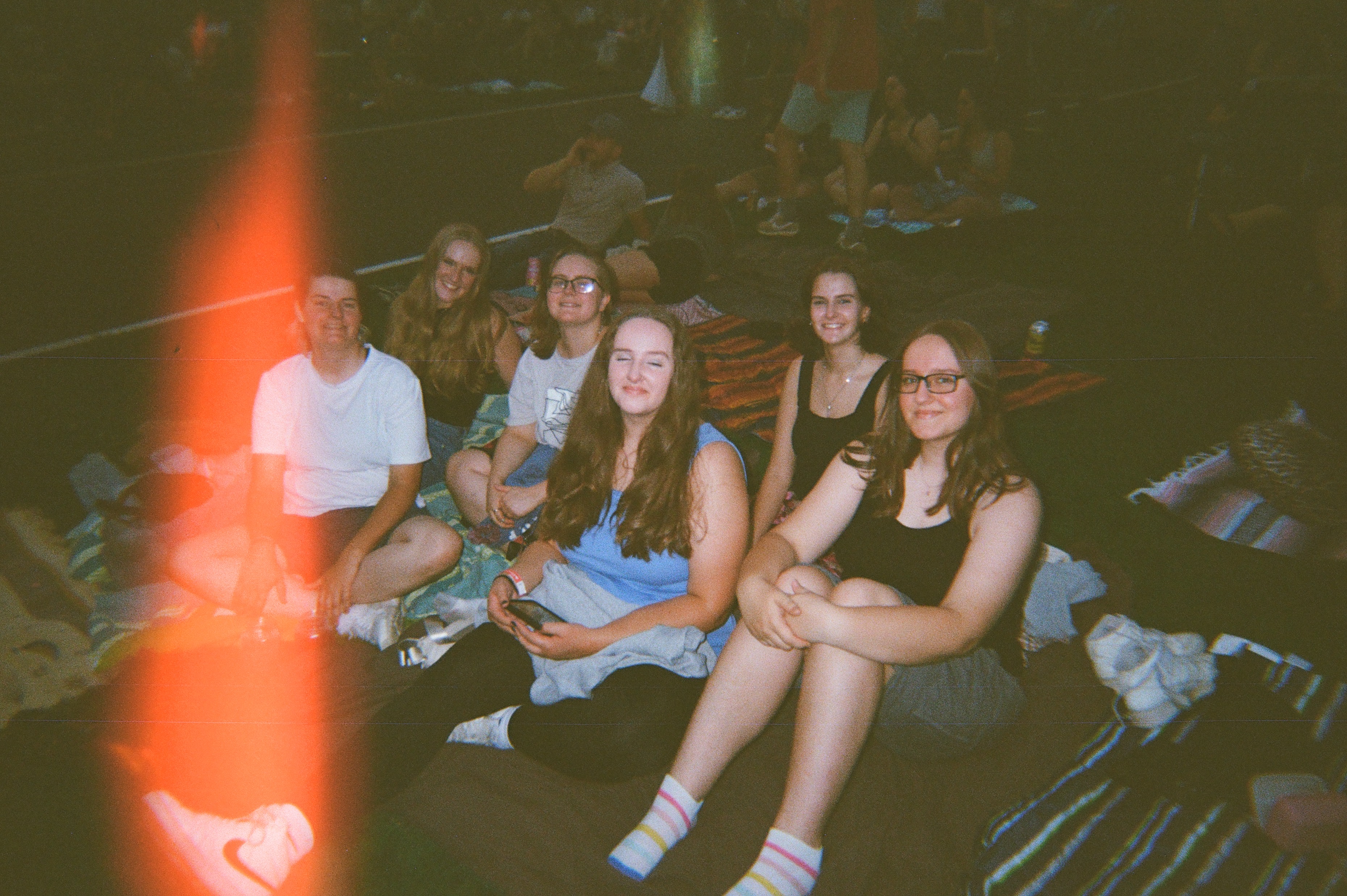 Six girls sitting on a blanket at a concert, light leak on the middle left.