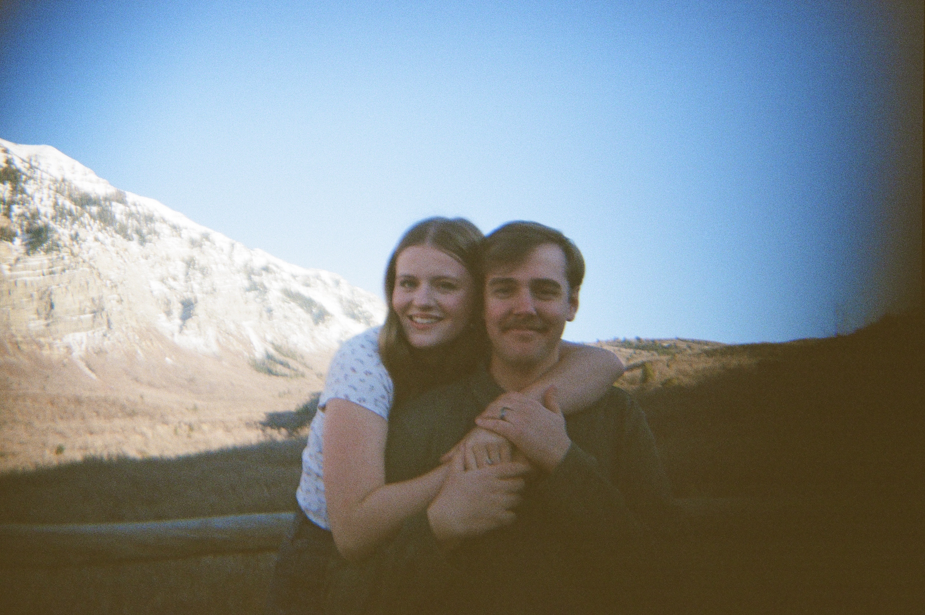 Young couple posed standing by a fence with a mountain background; woman standing behind man with arms wrapped around him.