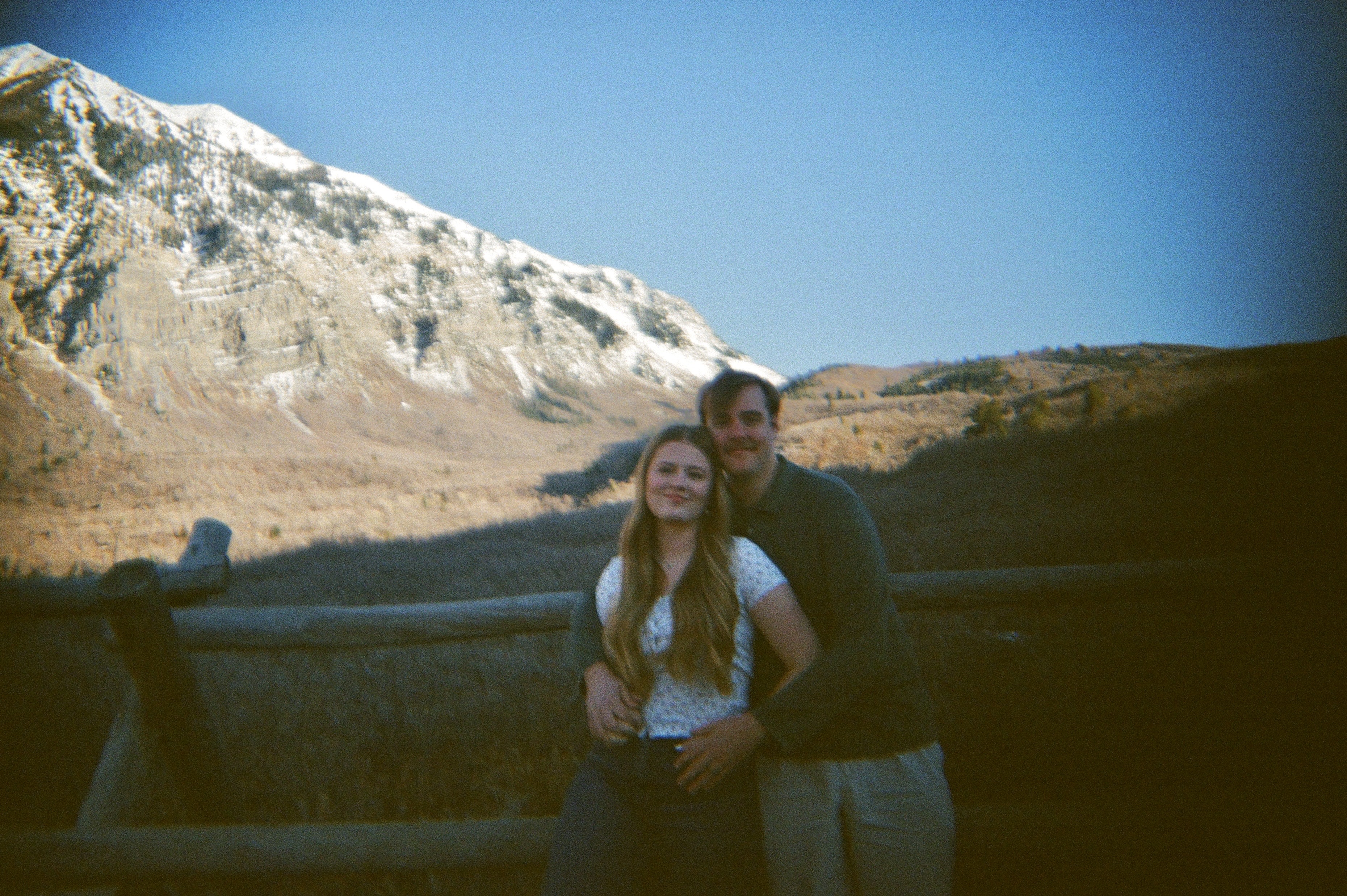 Young couple posed standing by a fence with a mountain background from a further distance.