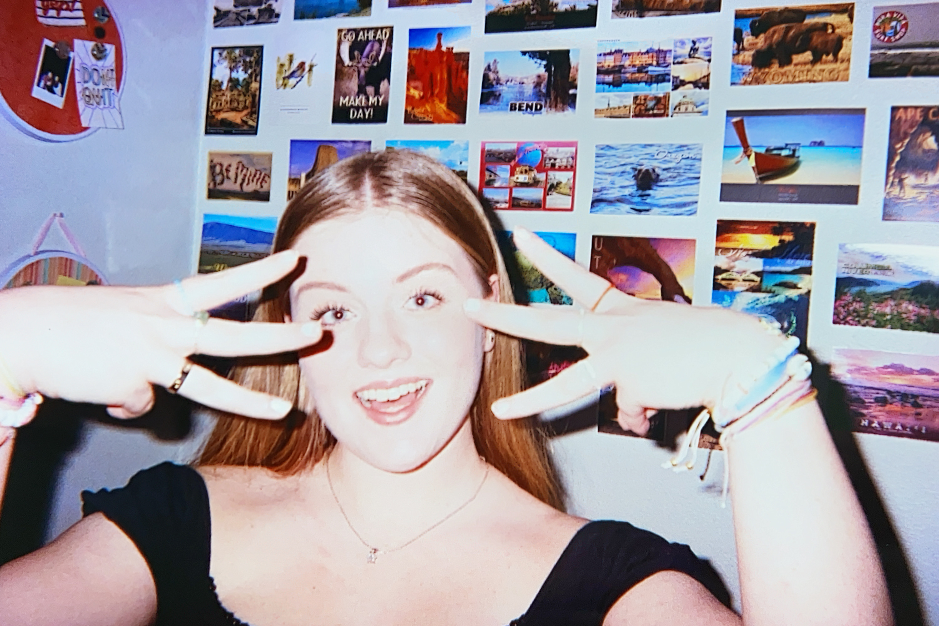 Young woman standing with her hands out in front of her face, wall of postcards in the background.
