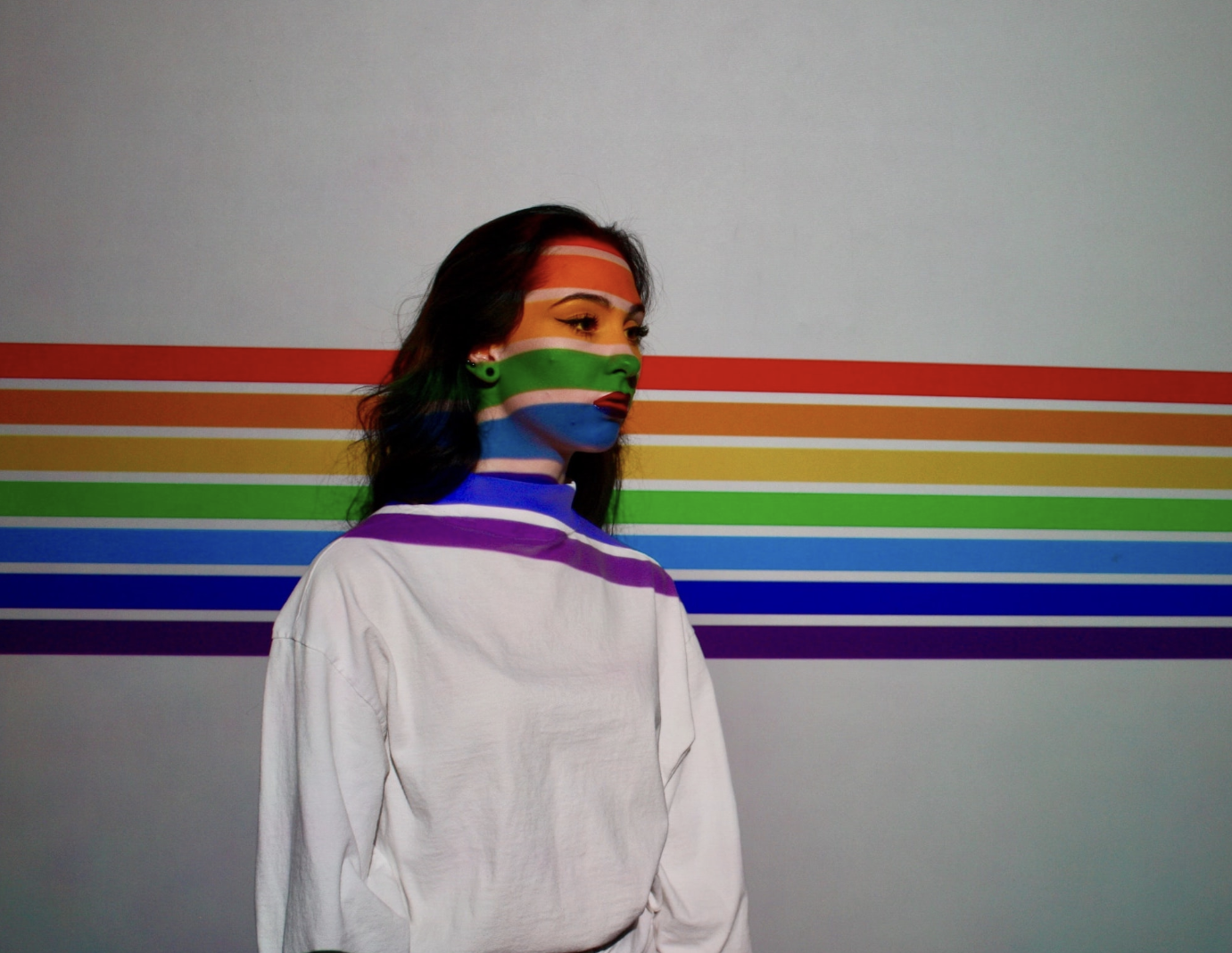 A person with curly brown hair and a white long-sleeve shirt looks to the side as the rainbow stripes of the pride flag are projected onto her face and the wall behind her.