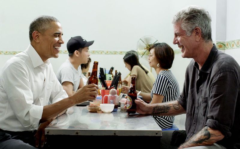 Bourdain and Obama sit in a crowded Vietnamese restaurant in NYC, sipping on beers and enjoying a meal together.