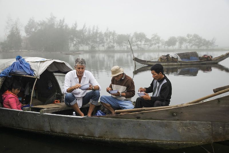 Bourdain sits in a small fishing boat in a port in Vietnam, eating a bowl of pho with local fishermen.