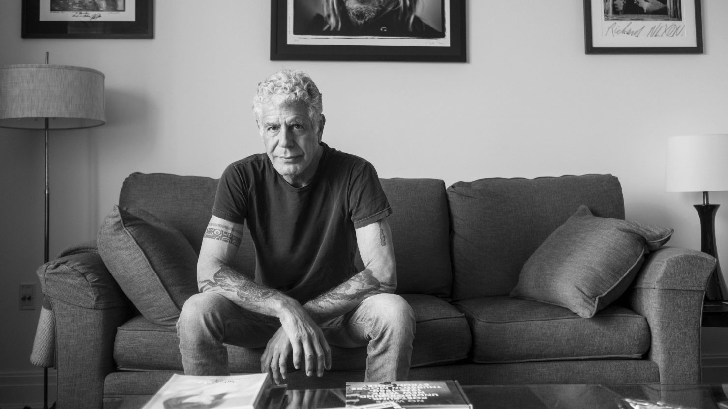 A grey haired Bourdain sits on a leather couch, leering at the camera. The photo is black and white.