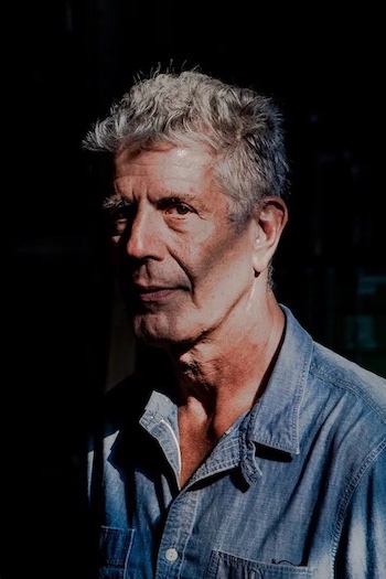 Elderly Bourdain looking smug in front of a black background. Shadows obsure his face.