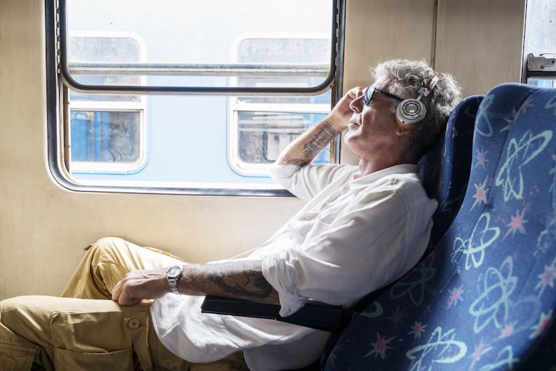 An older Bourdain sits on a train, sunglasses and headphones on.