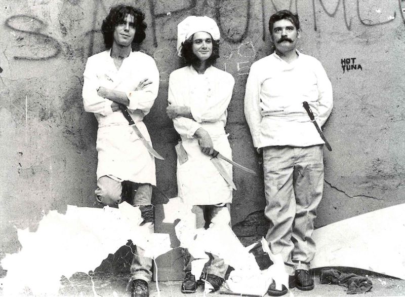 Teenage Bourdain standing against a brick wall in chef's whites. He stands next to two surly looking line cooks.'