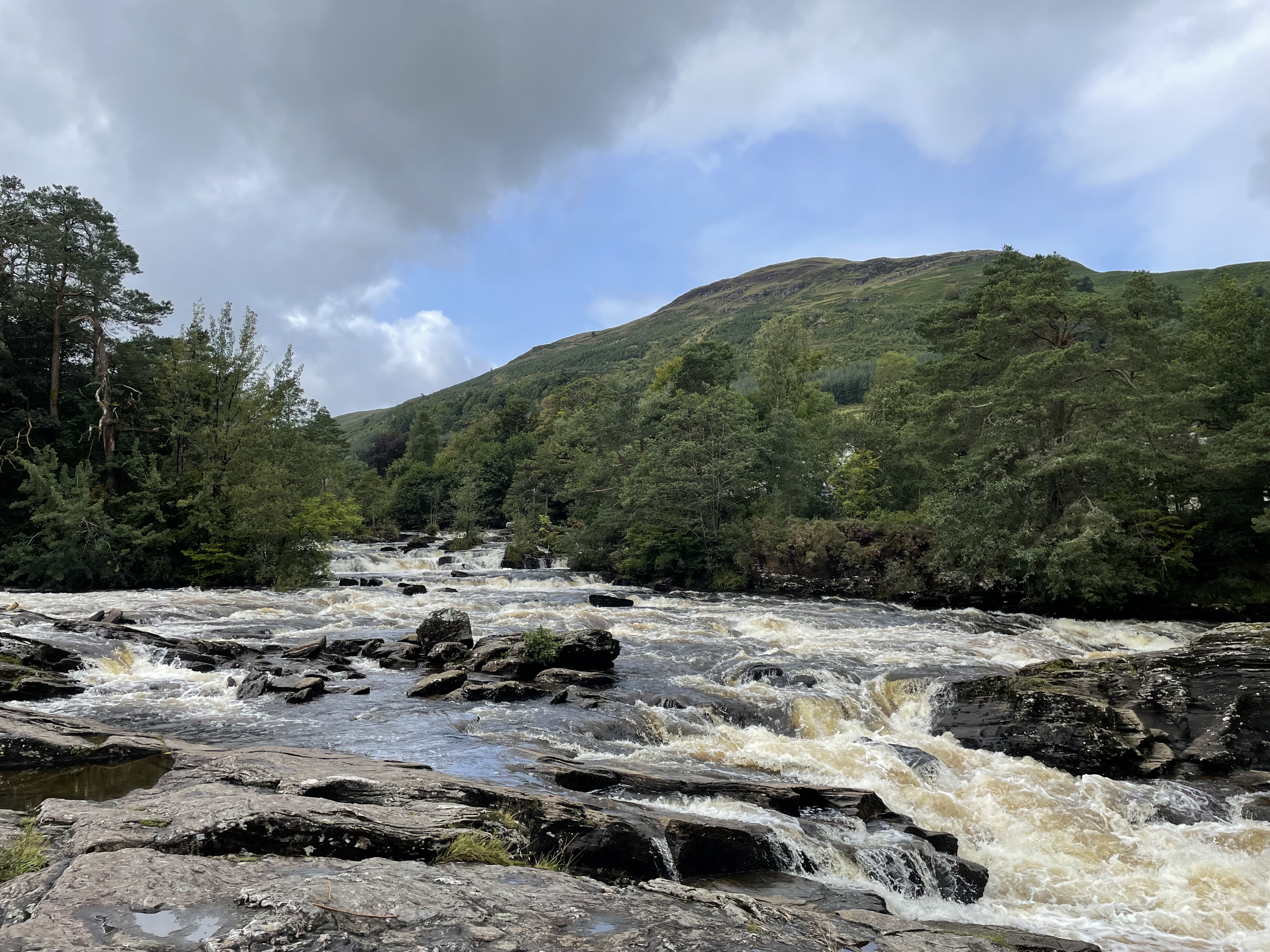 white waters rushing toward a waterfall set beside green hills and beneath a cloudy post-rain sky